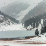 A realistic winter photograph inspired by Ratti Gali Lake in Pakistan, with snowfall covering the landscape. The scene shows the alpine lake surrounded with snow